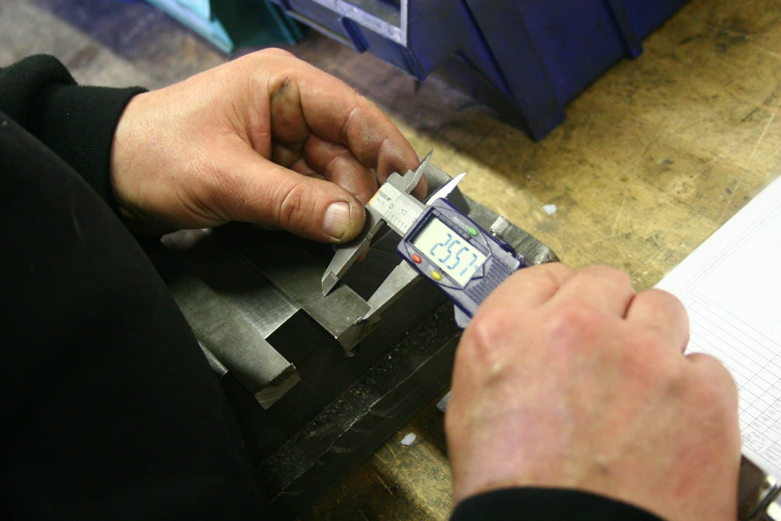 Technician measuring a machined part with a digital caliper beside an inspection sheet