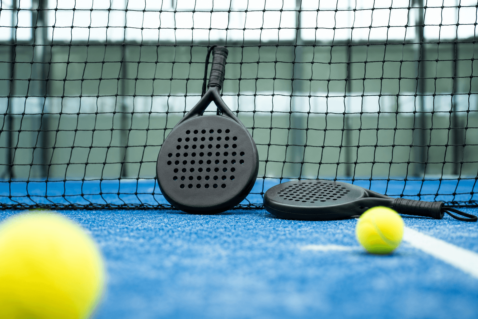 Two unbranded carbon padel rackets and balls resting against the net on a blue court